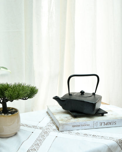 Black teapot on a stack of books with a plant and white curtain in the background