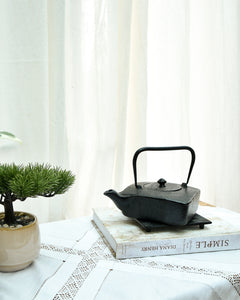 Black teapot on a stack of books with a plant and white curtain in the background